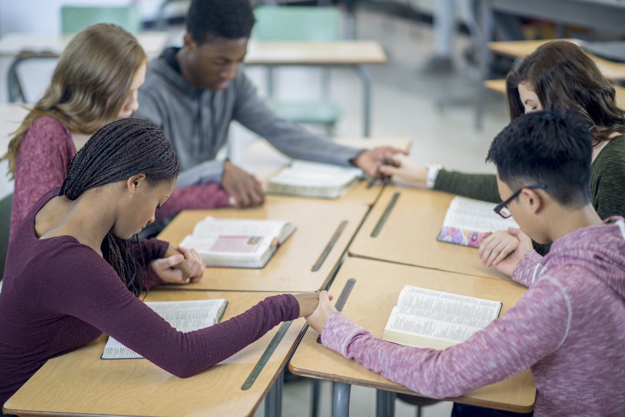 A multi-ethnic group of high school age students are sitting together at their desks and are holding hands while praying together.